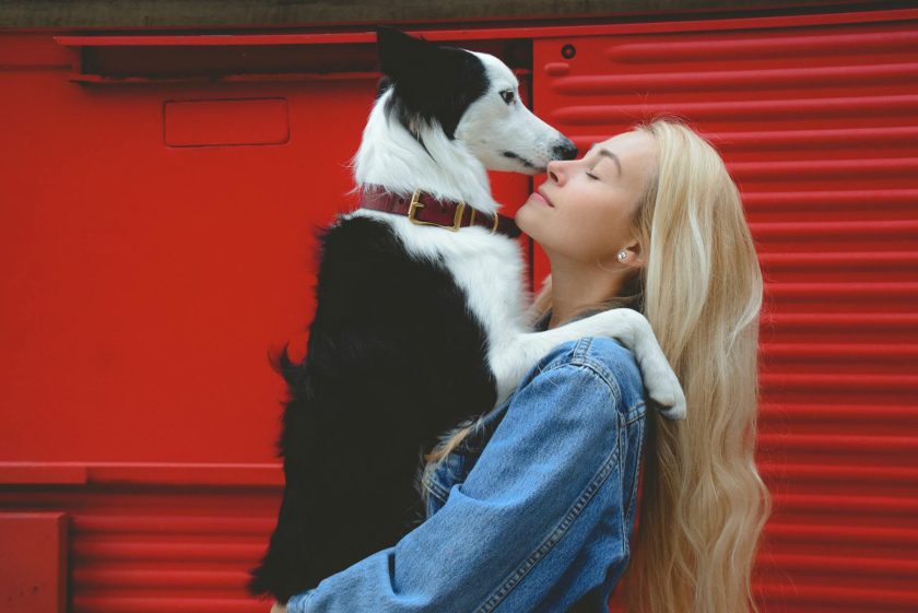 Blonde woman in denim jacket hugs black and white Border Collie dog against red metal shutter.