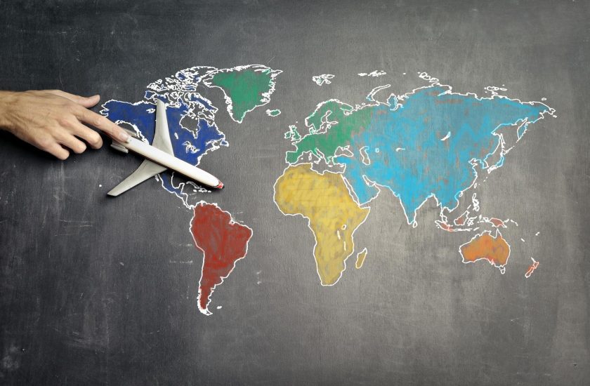 Hand holds white toy airplane over colorful chalk world map on blackboard.