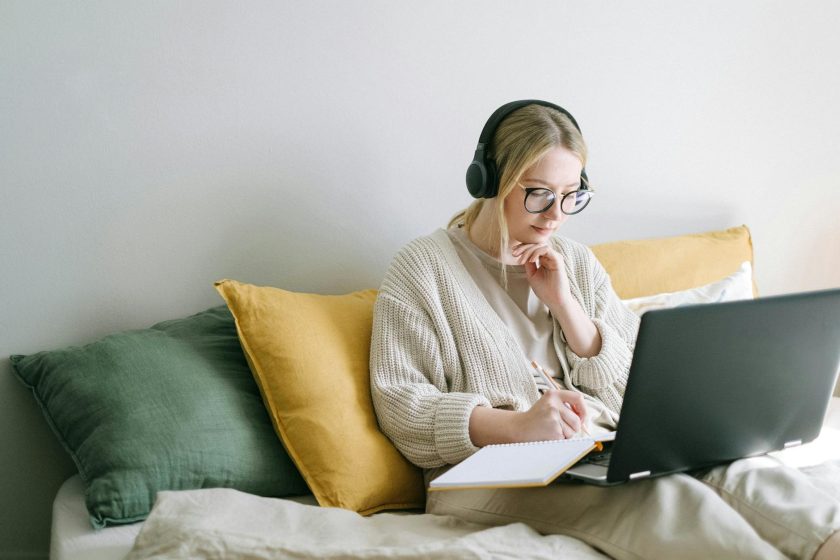 Woman with glasses and headphones sits on bed, types on laptop with notebook open.